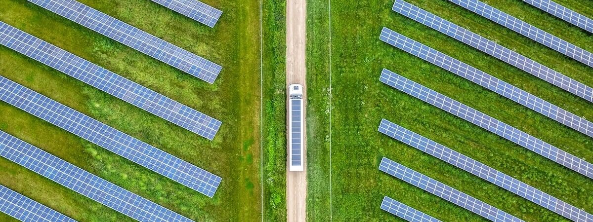 Aerial view of truck with solar on roof driving on light brown road between two green fields of blue solar panels, upwards through middle of image.