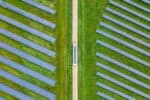 Aerial view of truck with solar on roof driving on light brown road between two green fields of blue solar panels, upwards through middle of image.