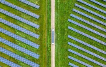 Aerial view of truck with solar on roof driving on light brown road between two green fields of blue solar panels, upwards through middle of image.