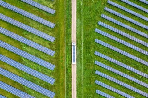 Aerial view of truck with solar on roof driving on light brown road between two green fields of blue solar panels, upwards through middle of image.