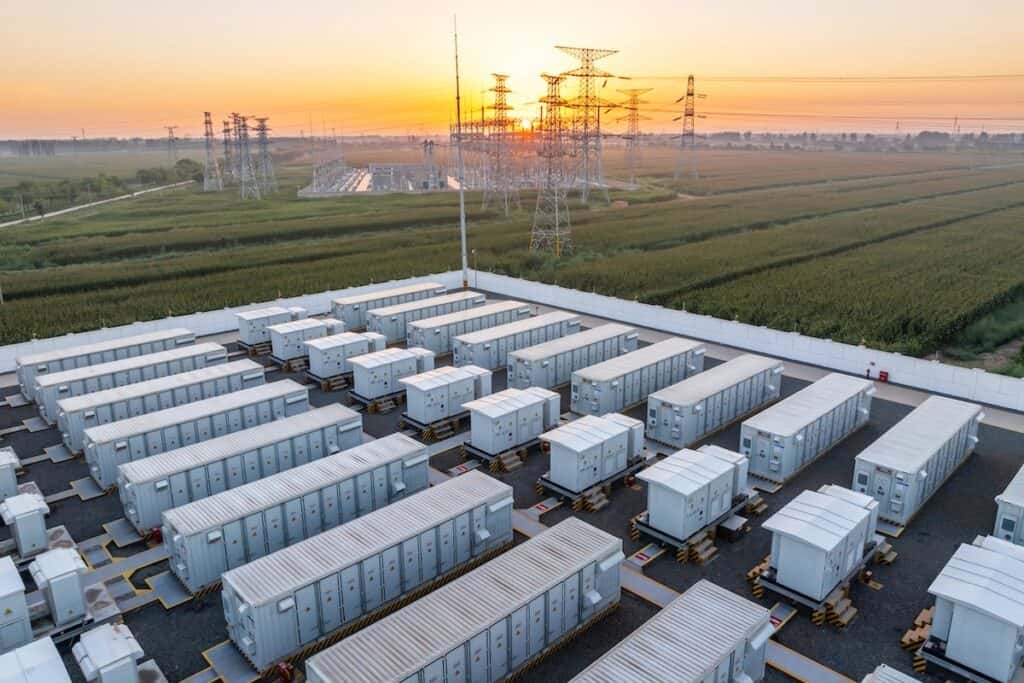 Energy storage power station at sunrise, with dozens of storage units in foreground, pylons and station infrastructure behind amongst green fields.