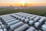 Energy storage power station at sunrise, with dozens of storage units in foreground, pylons and station infrastructure behind amongst green fields.
