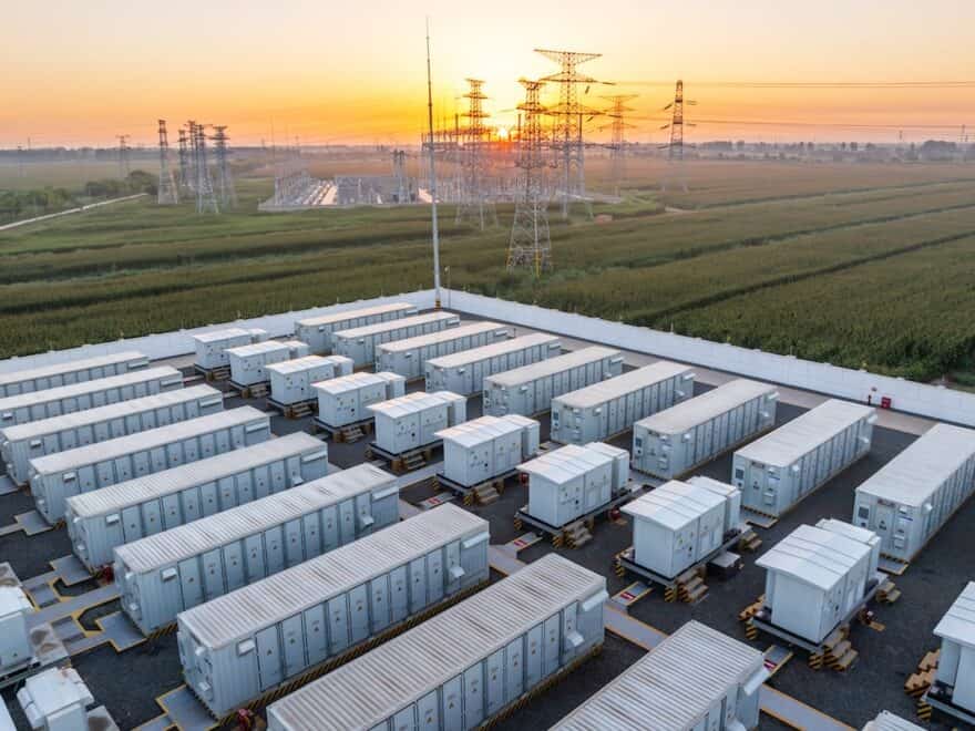 Energy storage power station at sunrise, with dozens of storage units in foreground, pylons and station infrastructure behind amongst green fields.