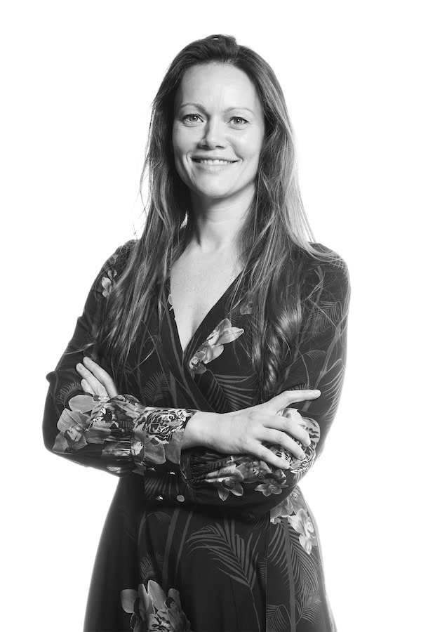 Black and white portrait of Kirstie David, pictured hair down and with arms crossed in long-sleeved dark dress with floral pattern, no background.