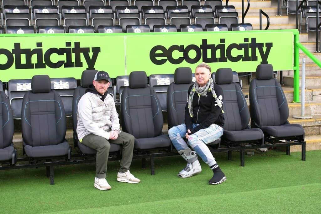 Rory MacFadyen of Reflo and Dale Vince of Forest Green Rovers seated in dugout at FGR stadium, with Ecotricity branding in black on green behind.
