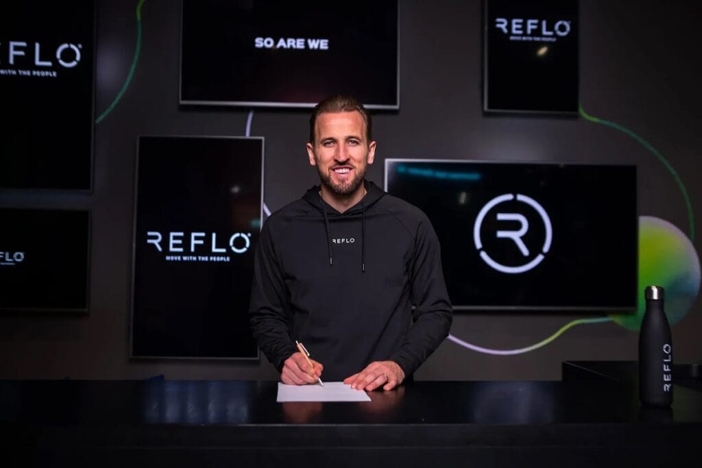 Footballer Harry Kane pictured in black Reflo hoodie signing paper at black desk infant of multiple screens displaying Reflo name and logo.