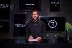 Footballer Harry Kane pictured in black Reflo hoodie signing paper at black desk infant of multiple screens displaying Reflo name and logo.