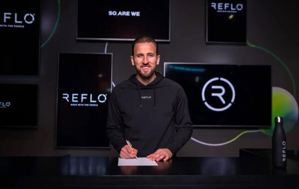 Footballer Harry Kane pictured in black Reflo hoodie signing paper at black desk infant of multiple screens displaying Reflo name and logo.