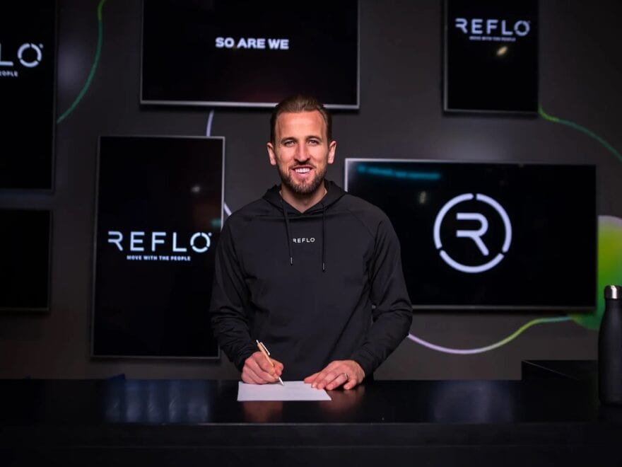 Footballer Harry Kane pictured in black Reflo hoodie signing paper at black desk infant of multiple screens displaying Reflo name and logo.