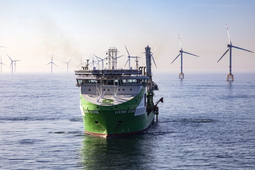 Ship seen sailing to camera, pictured out at sea in green and white DEME livery, with wind farm behind