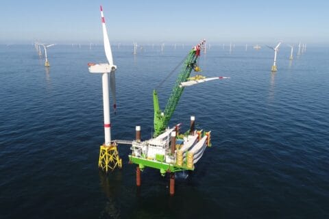 Neptune installation deck with crane shown at sea in green and white DEME livery, in position beside turbine with offshore wind farm behind.
