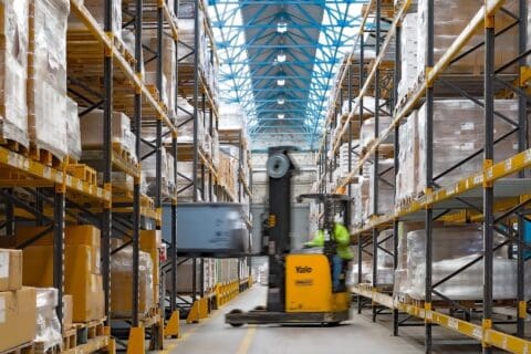 Yellow forklift blurred in action between warehouse racking loading Geopack tote box onto shelving.
