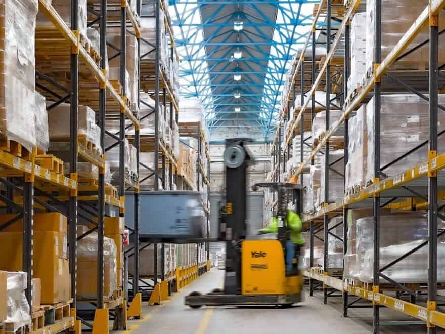 Yellow forklift blurred in action between warehouse racking loading Geopack tote box onto shelving.