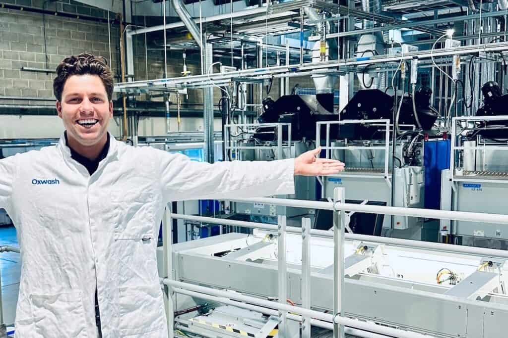 Oxwash Founder Kyle Grant pictured smiling in white workwear bearing company name, arms outstretched in front of laundry machinery.