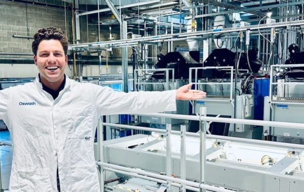 Oxwash Founder Kyle Grant pictured smiling in white workwear bearing company name, arms outstretched in front of laundry machinery.