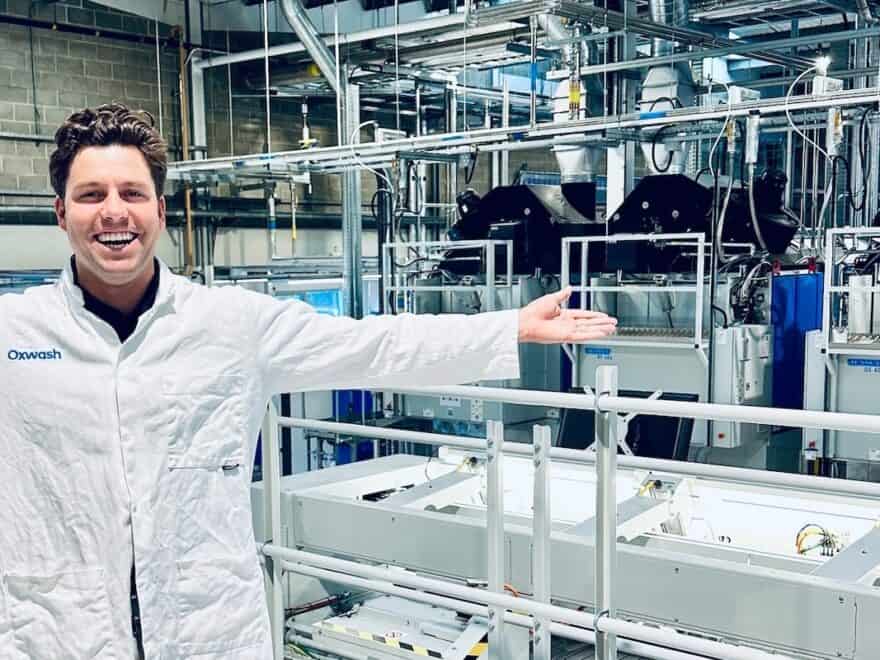 Oxwash Founder Kyle Grant pictured smiling in white workwear bearing company name, arms outstretched in front of laundry machinery.