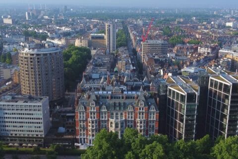Aerial cityscape of central London, green trees in foreground, Mandarin Oriental hotel front and centre, rotunda building to left and view down long straight street in the middle of the shot, receding into distance.