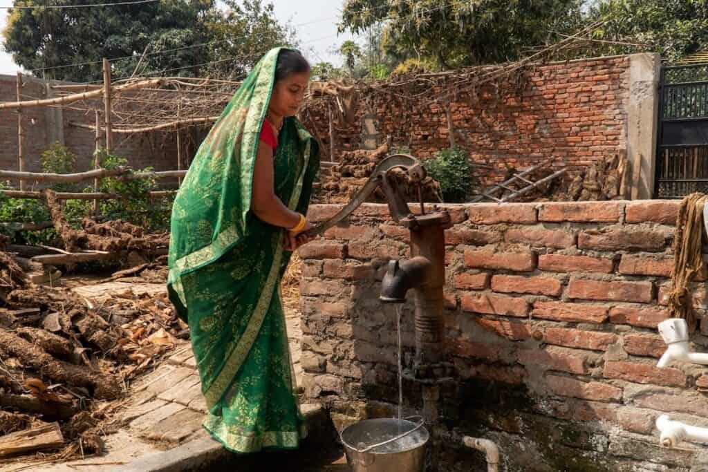 Indian female, in traditional light green sari, uses old metal hand pump to collect water from large standpipe in front of low rustic brick wall.