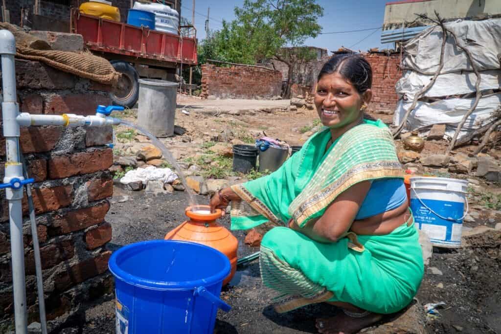 Indian female, smiling to camera in traditional light green sari, squats to collect water in orange pot from plastic pipe tap in left of shot.