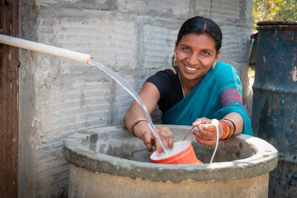 Indian female, smiling to camera in traditional blue sari, behind large stone well, collects water in orange pot from plastic pipe in foreground.