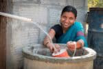 Indian female, smiling to camera in traditional blue sari, behind large stone well, collects water in orange pot from plastic pipe in foreground.