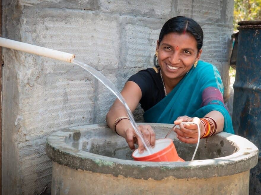 Indian female, smiling to camera in traditional blue sari, behind large stone well, collects water in orange pot from plastic pipe in foreground.