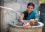 Indian female, smiling to camera in traditional blue sari, behind large stone well, collects water in orange pot from plastic pipe in foreground.