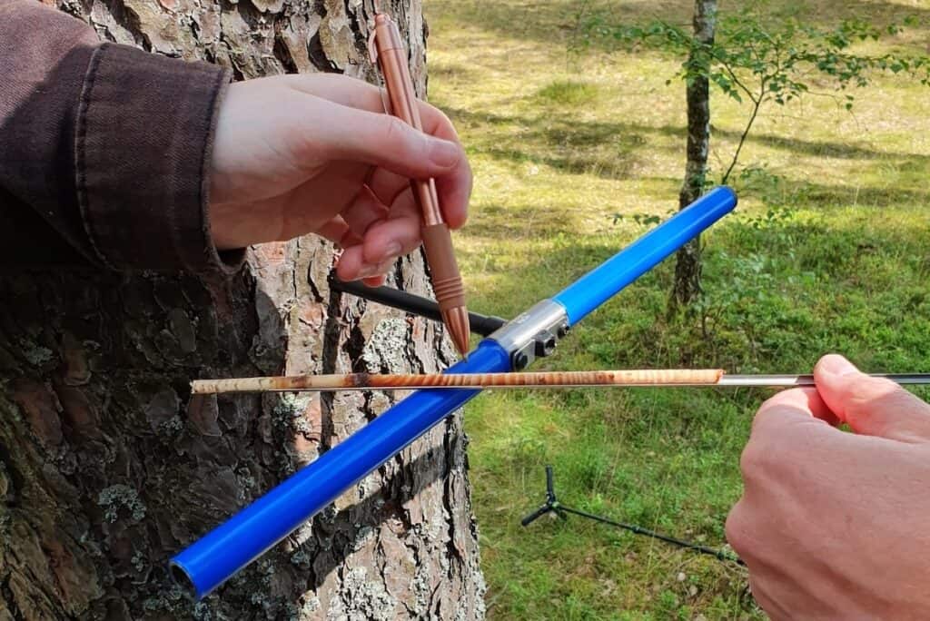 Close up of tree analysis shows hands in shot left and right, holding pen and core sample, with blue borer inserted into trunk between them.