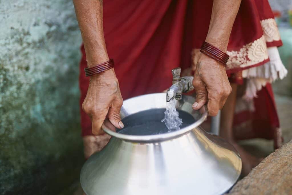 Close-up hands, with red bangles on bare arms, collect water in stainless steel container under metal tap, with red dress out of focus behind.