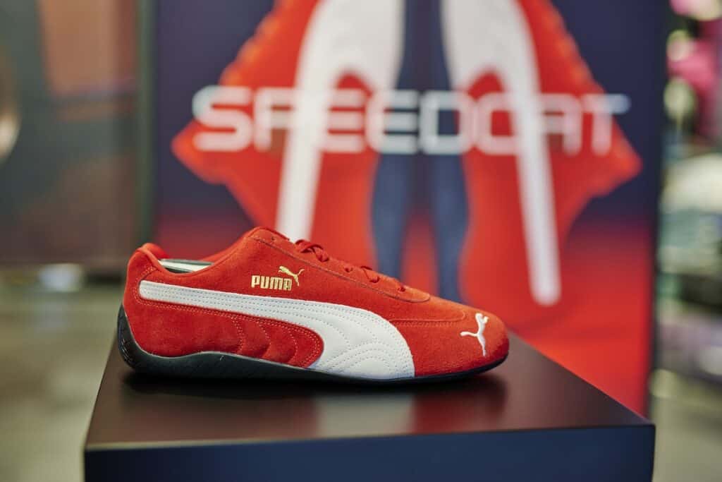 Close-up of red PUMA Speedcat shoe with white stripe and gold branding, pictured on plinth in store, with display signage blurred behind.