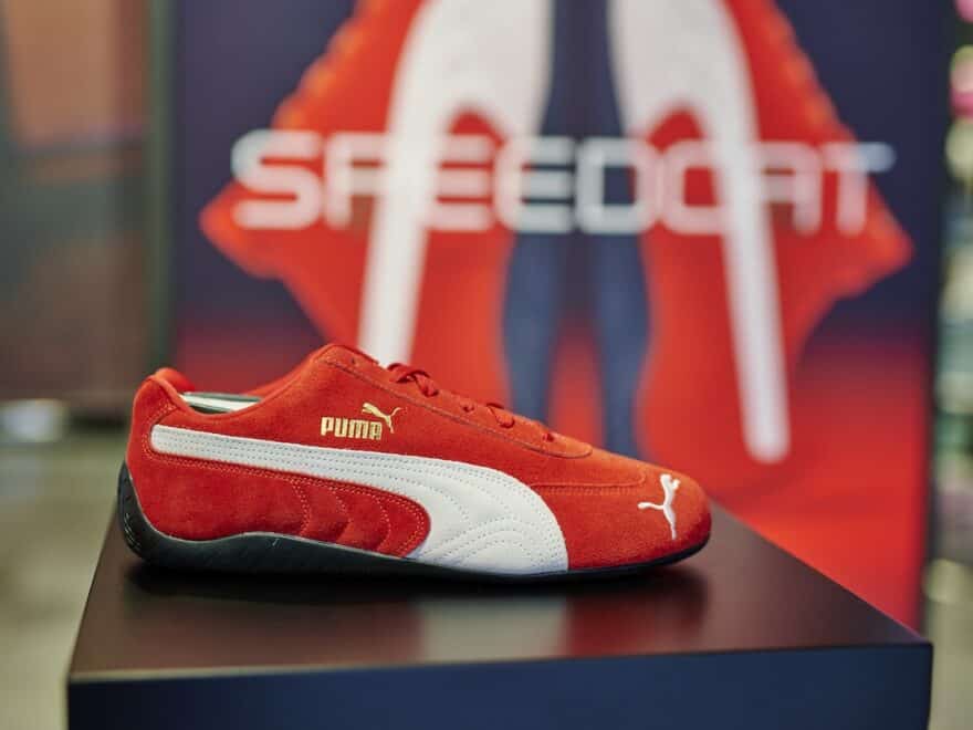 Close-up of red PUMA Speedcat shoe with white stripe and gold branding, pictured on plinth in store, with display signage blurred behind.