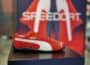 Close-up of red PUMA Speedcat shoe with white stripe and gold branding, pictured on plinth in store, with display signage blurred behind.