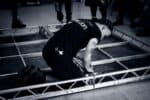 Black and white image of male stagehand in branded Connection Crew t-shirt, knelt on floor, working within structural frame on reverse of panel.