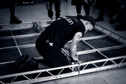 Black and white image of male stagehand in branded Connection Crew t-shirt, knelt on floor, working within structural frame on reverse of panel.