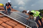 Two workers in high-vis and white hard hats are shown installing solar panels on terracotta tiles of residential roof.