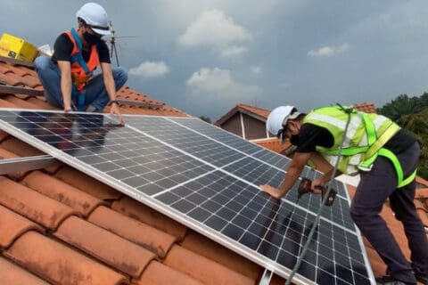 Two workers in high-vis and white hard hats are shown installing solar panels on terracotta tiles of residential roof.