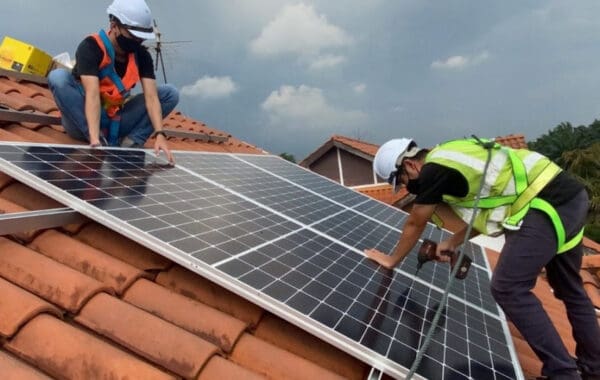 Two workers in high-vis and white hard hats are shown installing solar panels on terracotta tiles of residential roof.