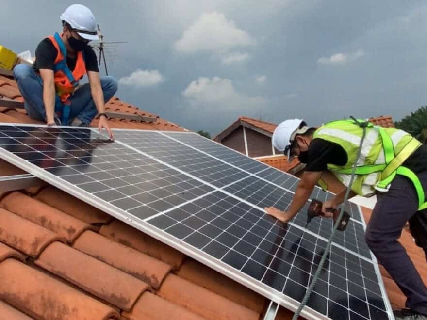 Two workers in high-vis and white hard hats are shown installing solar panels on terracotta tiles of residential roof.