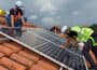 Two workers in high-vis and white hard hats are shown installing solar panels on terracotta tiles of residential roof.