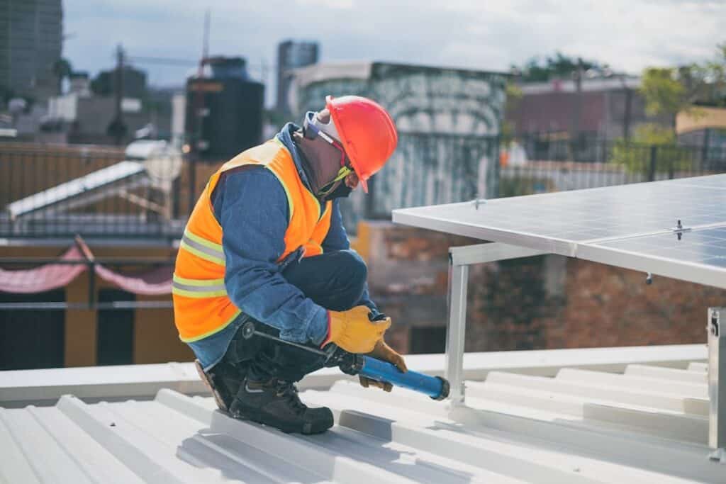 Close-up image shows person in orange high-vis and red hard hat working on installation of rooftop solar panels, with urban landscape in background.