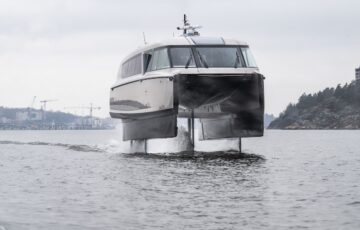 Candela P-12, the world's first electric hydrofoil ferry, pictured head-on, in full flow and up out of the water, coastline in background.