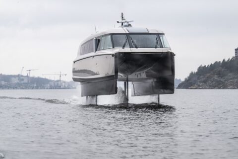Candela P-12, the world's first electric hydrofoil ferry, pictured head-on, in full flow and up out of the water, coastline in background.