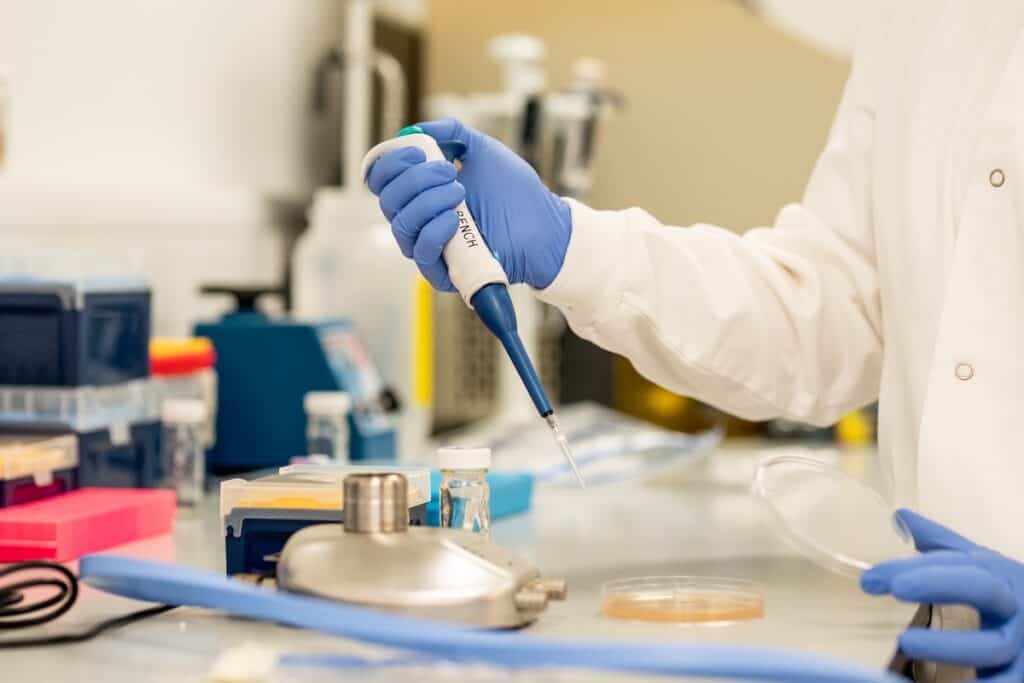 Close-up of person in white coat and blue surgical gloves using dropper to apply sample to Petri dish in laboratory setting.