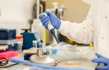 Close-up of person in white coat and blue surgical gloves using dropper to apply sample to Petri dish in laboratory setting.