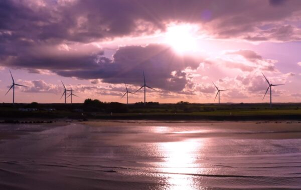 Onshore wind turbines pictured at sunset by water's edge.