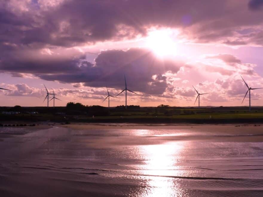 Onshore wind turbines pictured at sunset by water's edge.