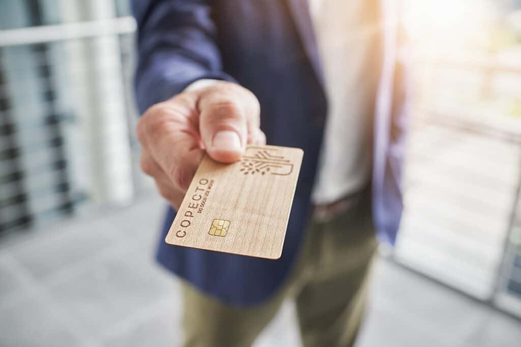 Wood COPECTO bank card in foreground, presented chip first to viewer by male hand, purple jacket, green trousers, white shirt out of focus behind.