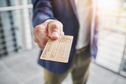 Wood COPECTO bank card in foreground, presented chip first to viewer by male hand, purple jacket, green trousers, white shirt out of focus behind.