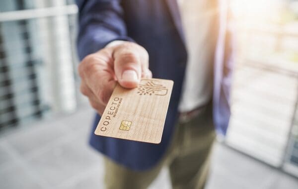 Wood COPECTO bank card in foreground, presented chip first to viewer by male hand, purple jacket, green trousers, white shirt out of focus behind.