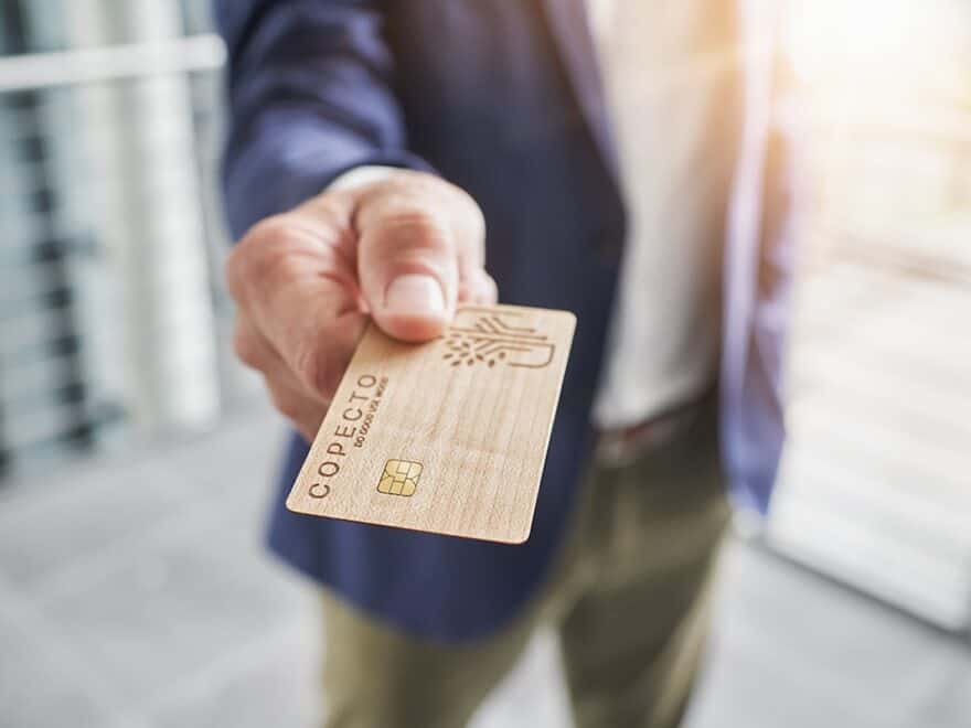 Wood COPECTO bank card in foreground, presented chip first to viewer by male hand, purple jacket, green trousers, white shirt out of focus behind.